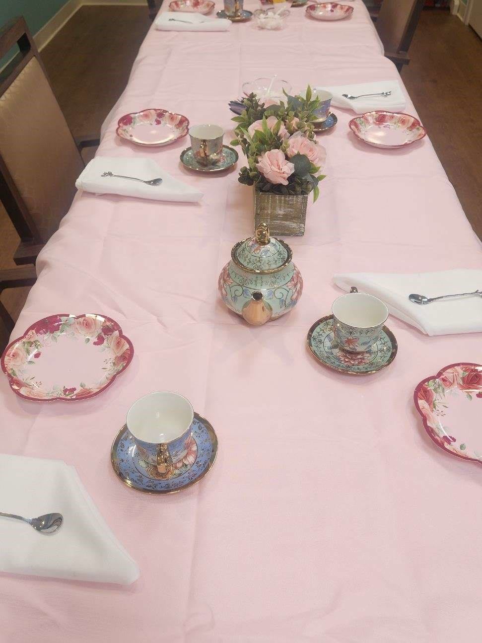 A beautifully arranged tea party setup with colorful teacups, plates, and a floral centerpiece on a pink tablecloth.