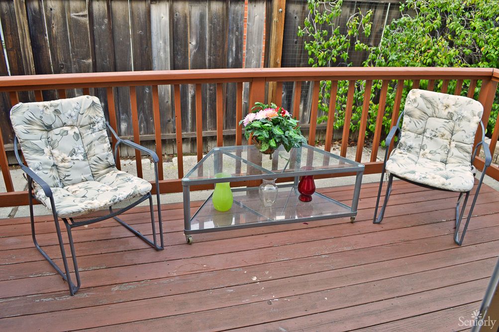 An outdoor patio with a glass table, two chairs, and a potted plant on a wooden deck.