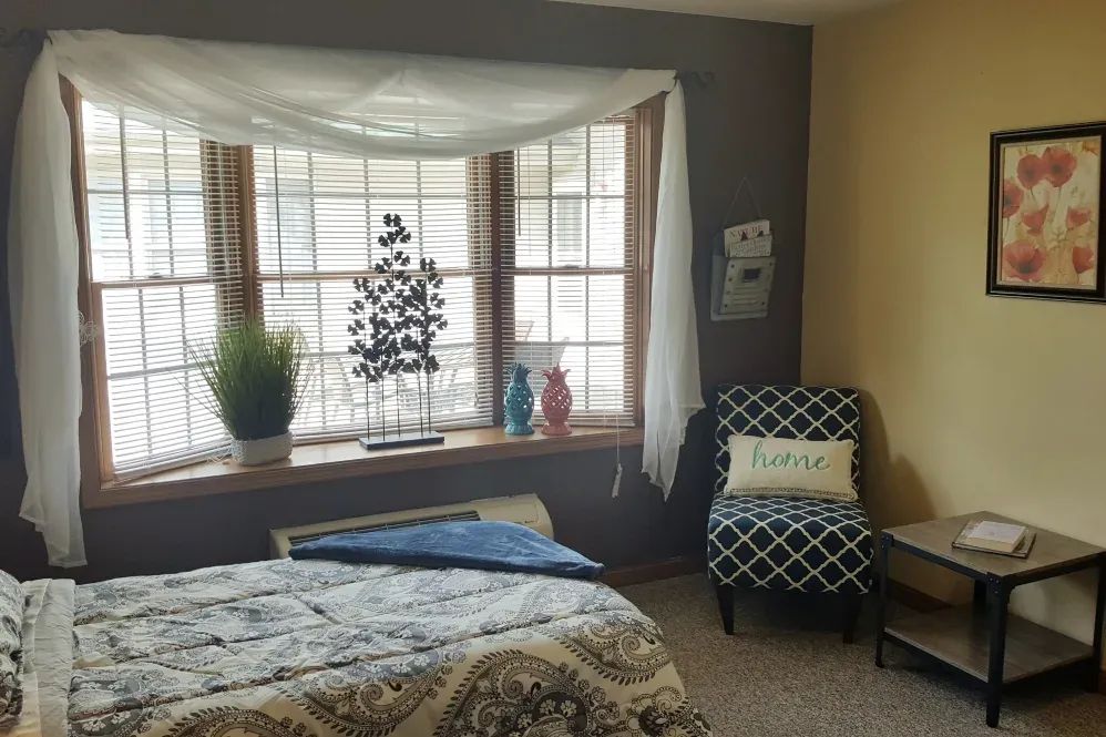 A cozy bedroom with a large window, patterned chair, and decorative plants.
