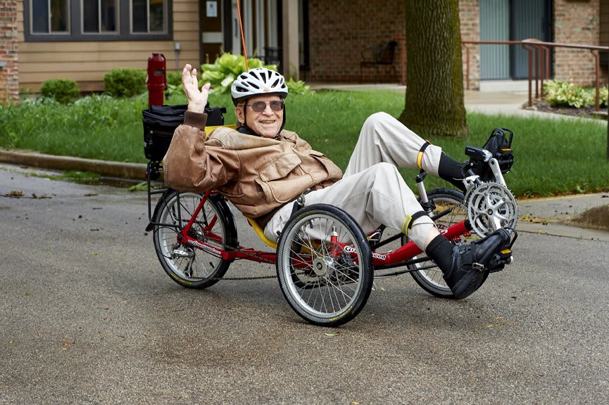 A person wearing a helmet is smiling and waving while riding a tricycle outdoors.