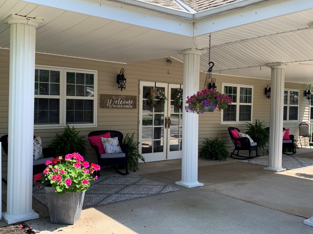The entrance of a building featuring a porch with seating and flower arrangements.