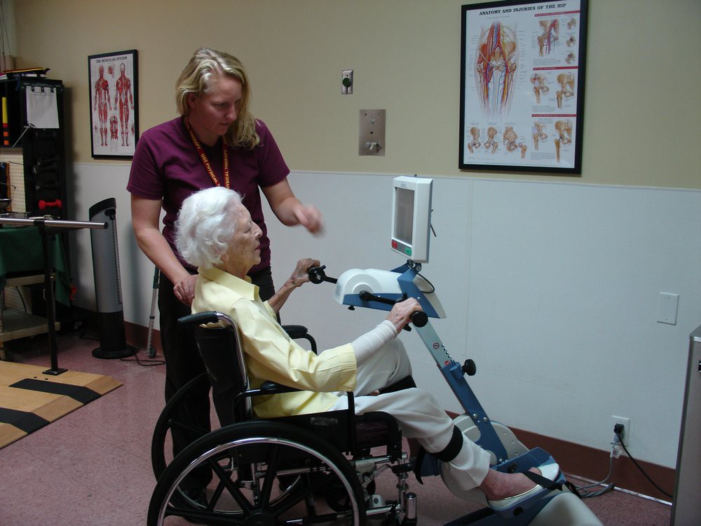 An elderly person in a wheelchair using exercise equipment with assistance from a caregiver.