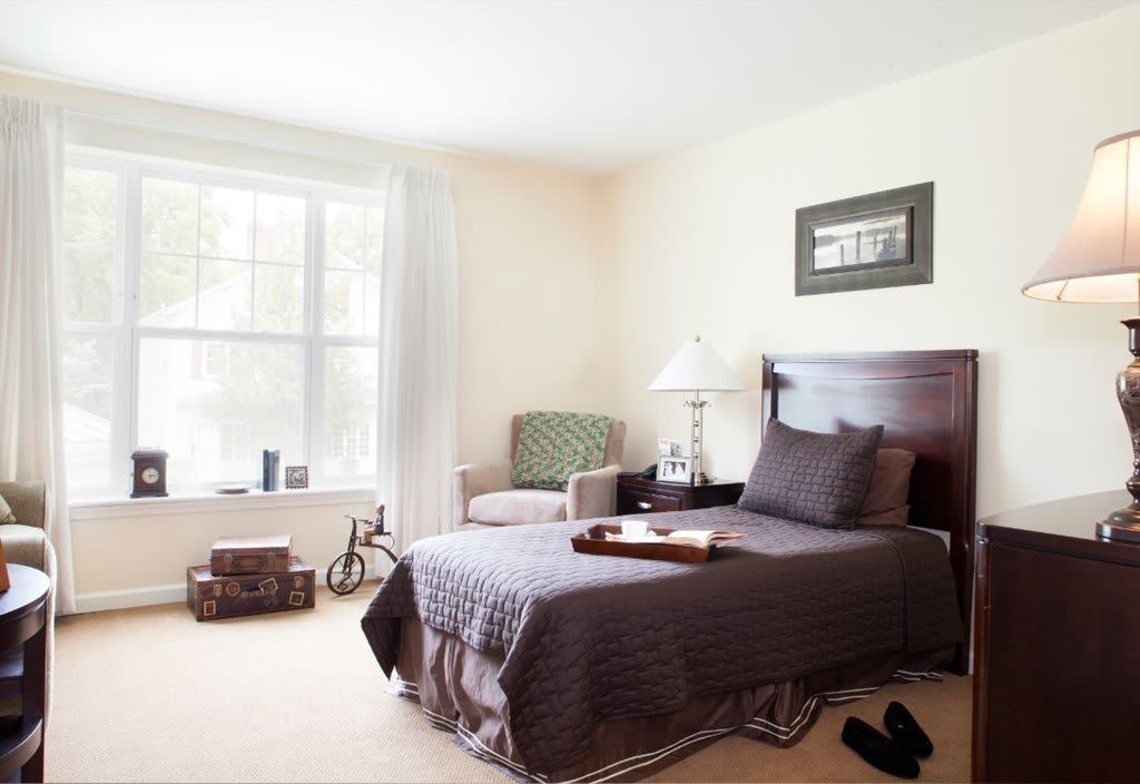A cozy bedroom with a large window, featuring a neatly made bed, a chair, and decorative elements.