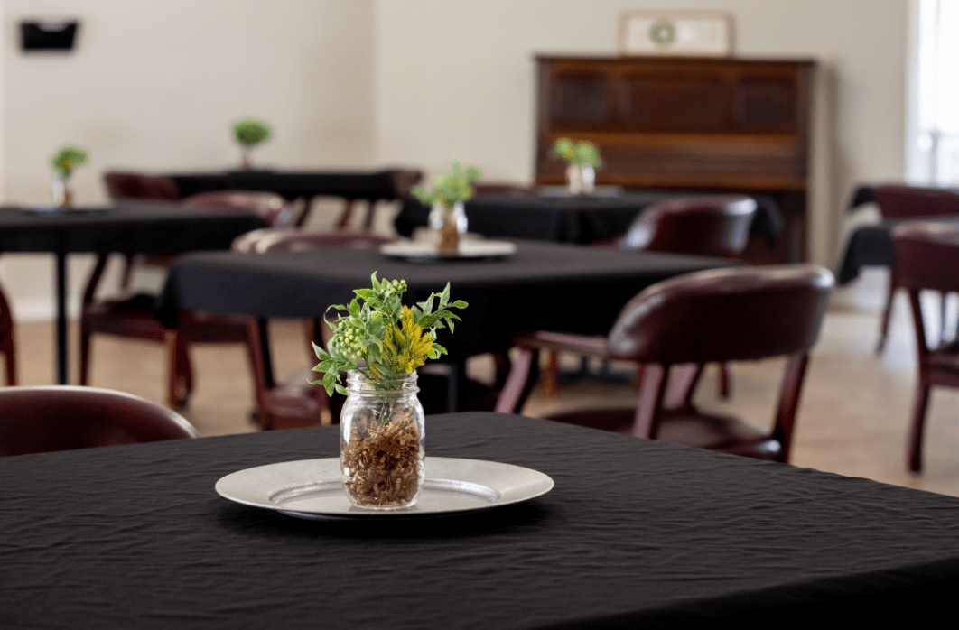 A dining area with tables covered in black cloth, each adorned with a jar centerpiece containing greenery.