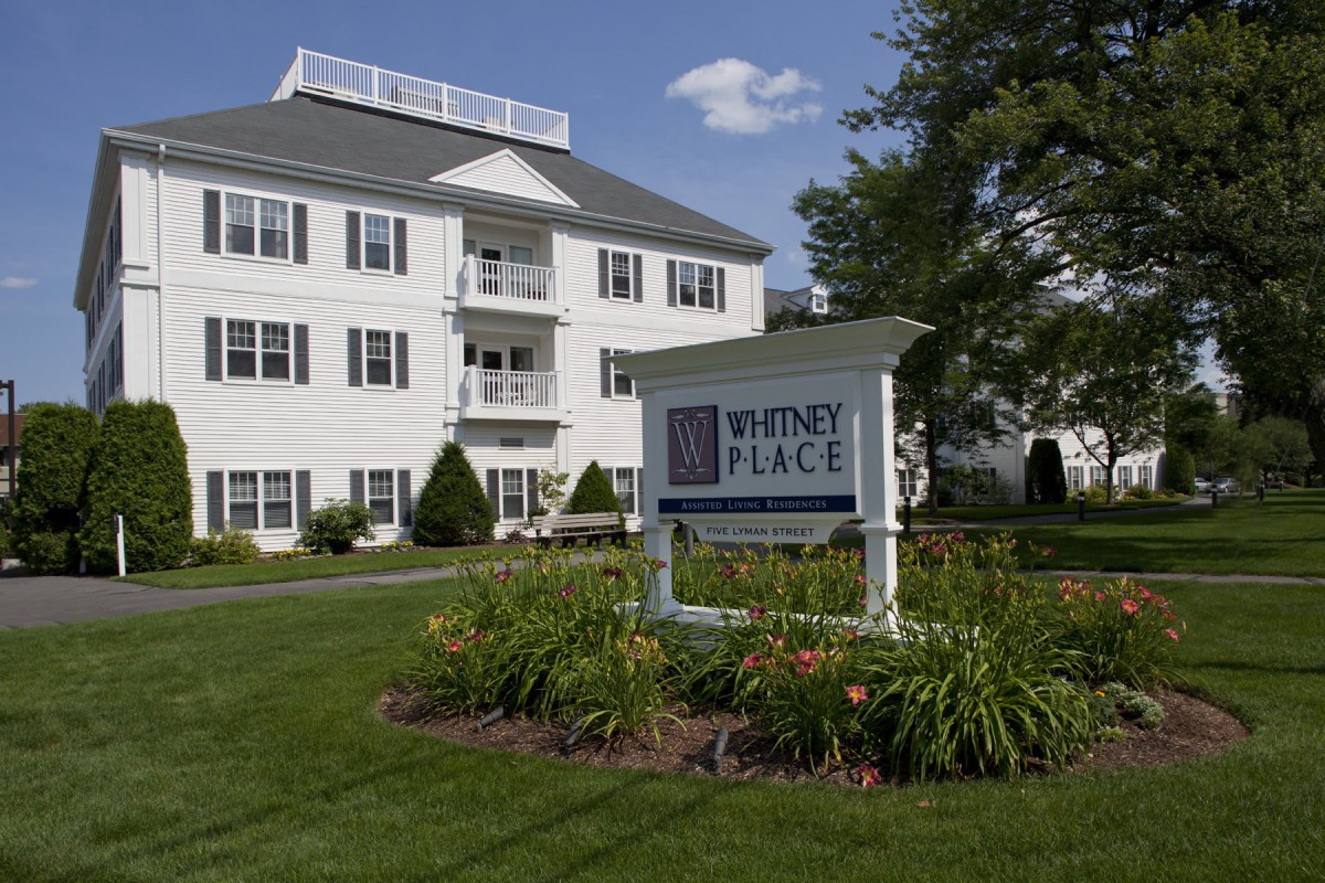 A white residential building with a sign labeled 'Whitney Place' surrounded by well-maintained landscaping and flowers.