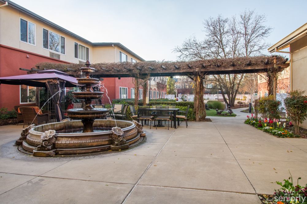 A tranquil courtyard featuring a central fountain, surrounded by outdoor seating and a pergola, with garden views and an adjacent building.