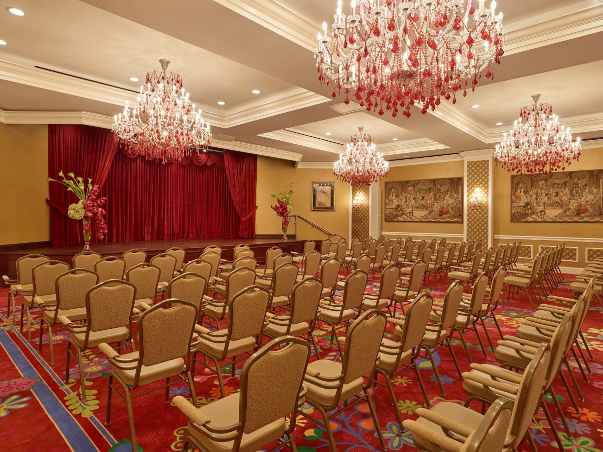 An elegant conference room with rows of chairs facing a stage, adorned with chandeliers and red curtains.