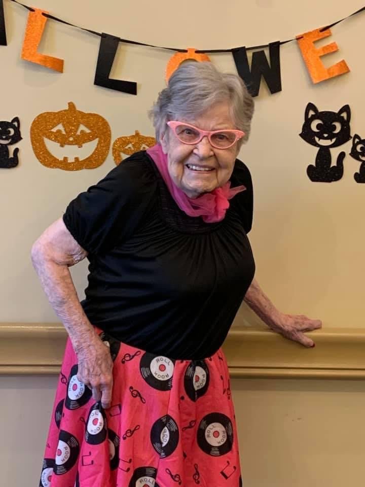 An elderly woman wearing a festive Halloween costume stands in front of Halloween-themed decorations.