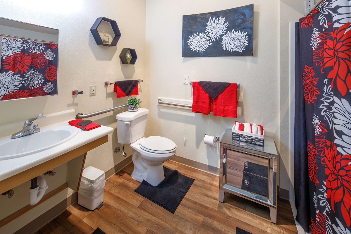 A modern bathroom with red and black towel accents, featuring a toilet, sink, and wooden flooring.