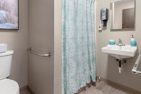 A bathroom featuring a shower with a patterned curtain, a sink with a mirror above it, and a soap dispenser on the wall.