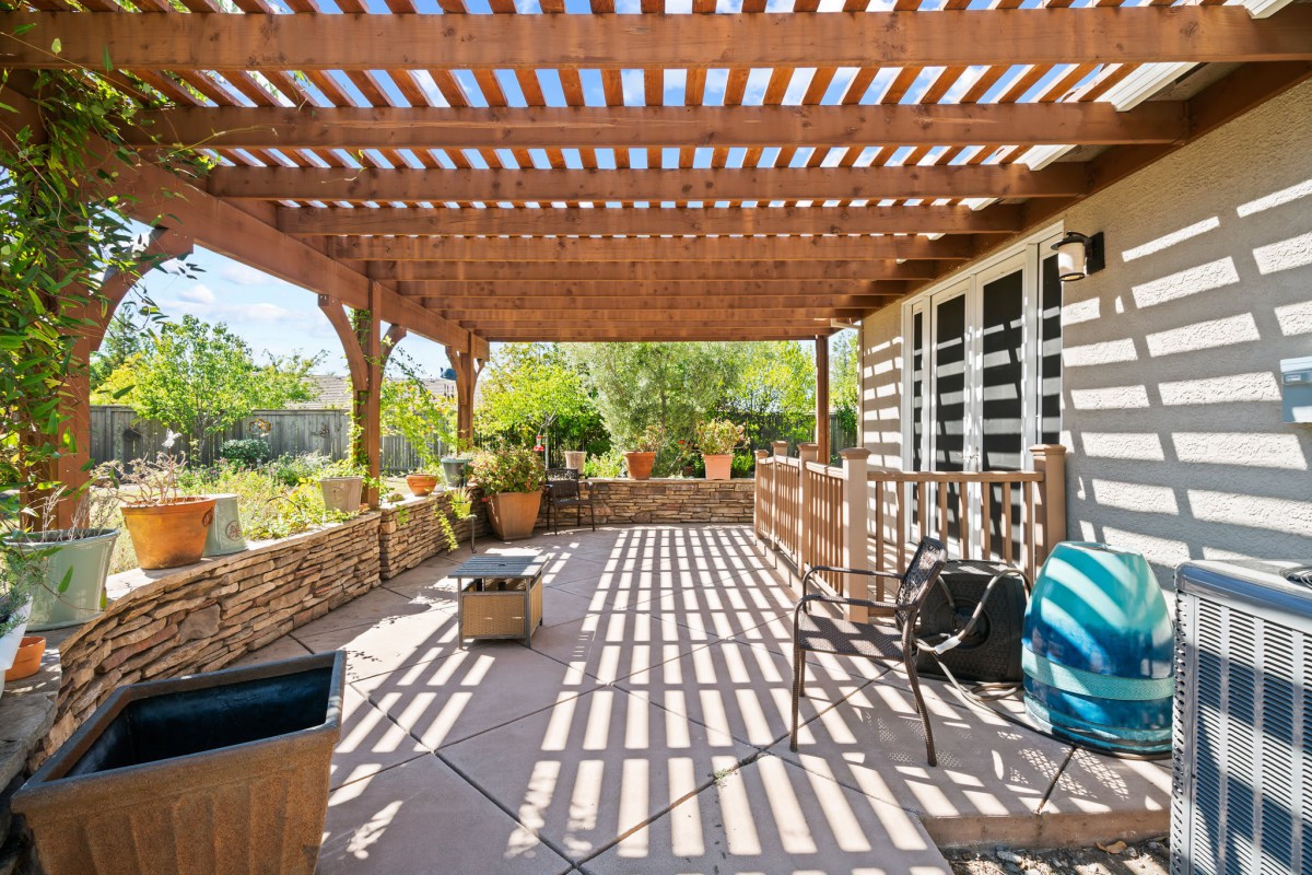 A sunny patio with a wooden pergola casting shadows, surrounded by garden plants and outdoor furniture.