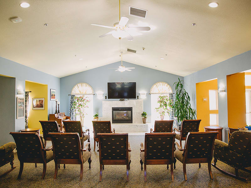 A spacious living room with arranged chairs facing a television above a fireplace, complemented by large windows and indoor plants.