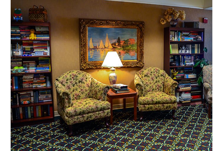 A cozy living room area with two armchairs, a lamp on a side table, bookshelves, and a painting on the wall.