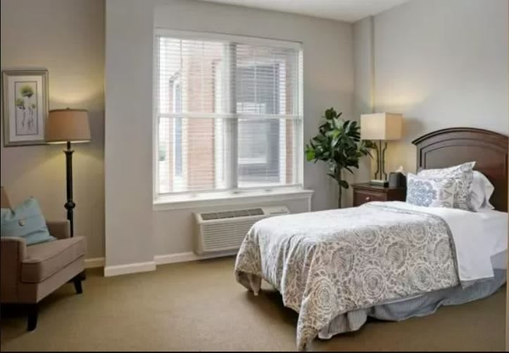 A cozy bedroom with neutral colors, featuring a bed, bedside lamps, a large window, and a potted plant.