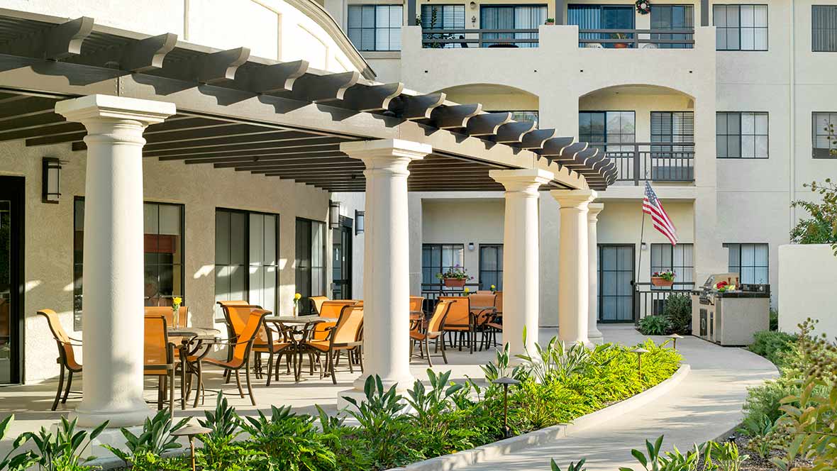 A charming outdoor seating area with wooden chairs and tables under a pergola, surrounded by lush greenery and elegant columns.