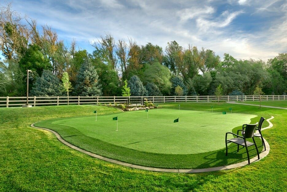 A well-maintained outdoor putting green surrounded by trees and a white fence, with a bench nearby.