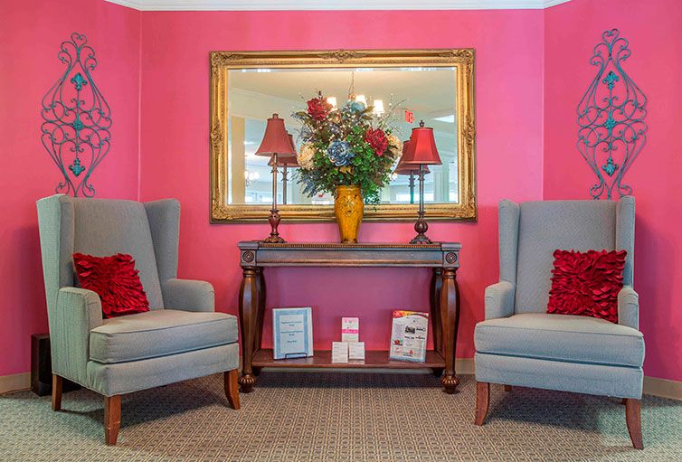 A cozy living area featuring pink walls, two gray armchairs, a decorative console table with a vase of flowers and lamps, and a large mirror.