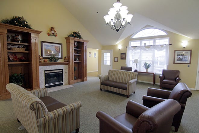 A cozy living room with a fireplace, sofas, and armchairs under a chandelier, featuring large windows for natural light.