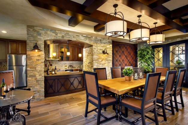 A modern dining room featuring a wooden table and chandeliers, with an open kitchen and stone wall.