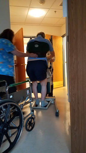 A healthcare worker assists a patient in a transfer using a mobility aid in a hospital setting.