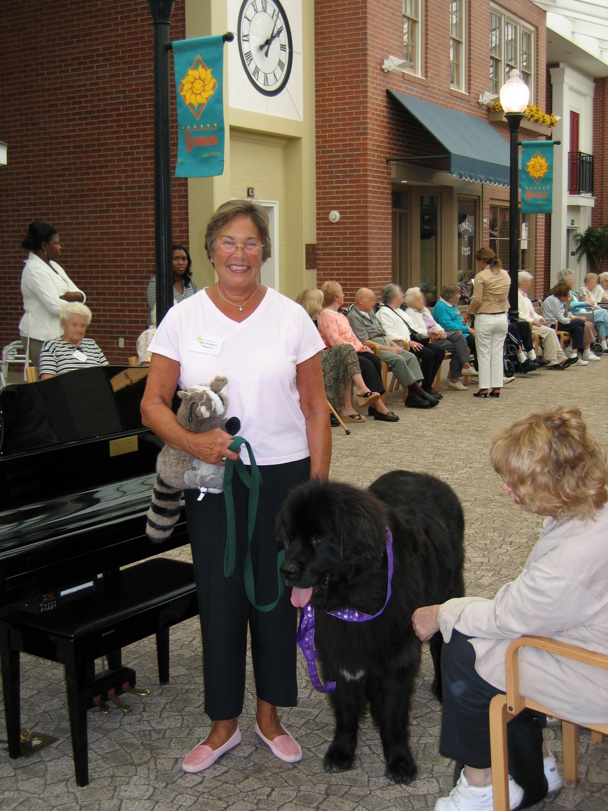 A person stands with a dog on a street while another person sits nearby, with a crowd in the background.