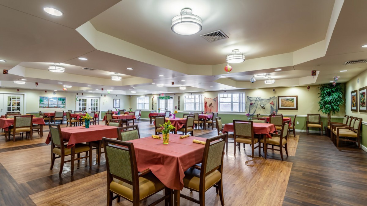 A brightly lit dining room with neatly arranged tables and chairs, featuring modern light fixtures and large windows.
