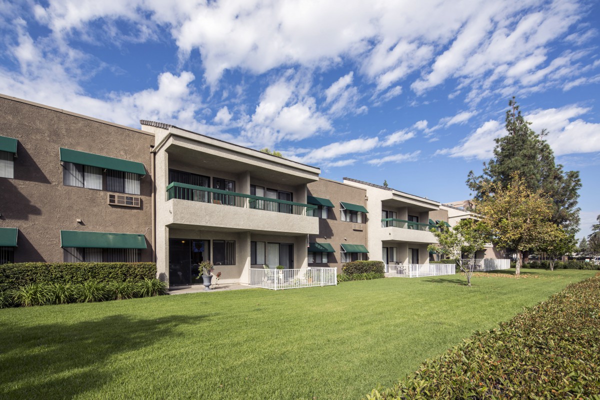 A modern apartment building with balconies, surrounded by a green lawn and trees under a blue sky with clouds.