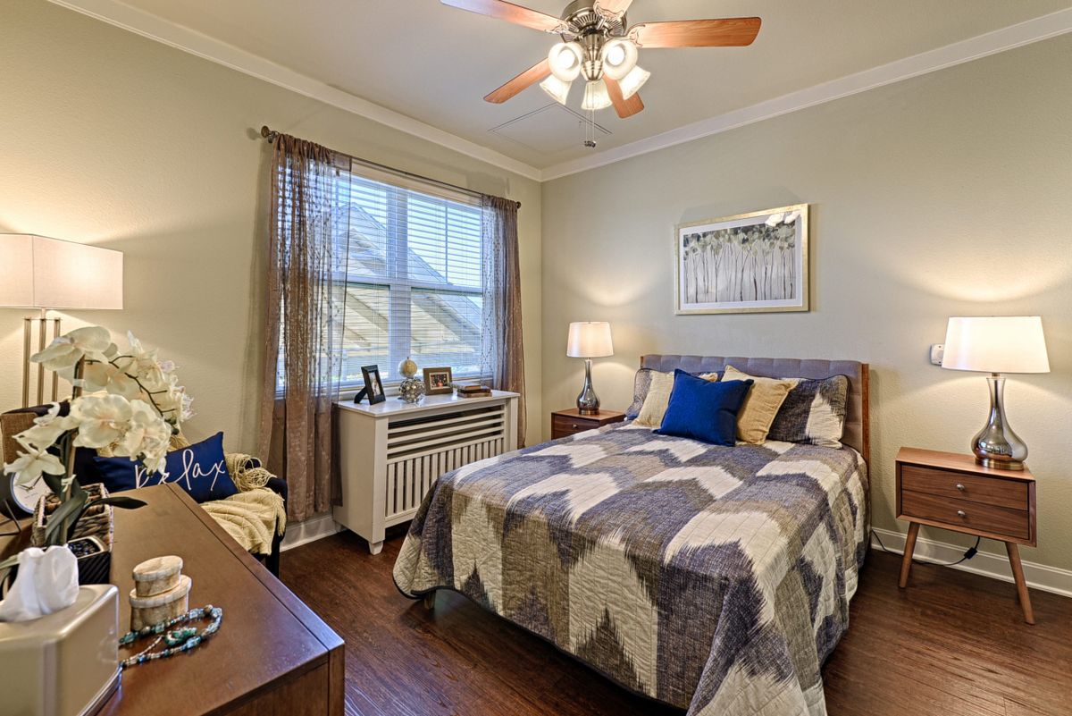 A cozy and well-decorated bedroom with a patterned quilt, ceiling fan, and natural lighting.