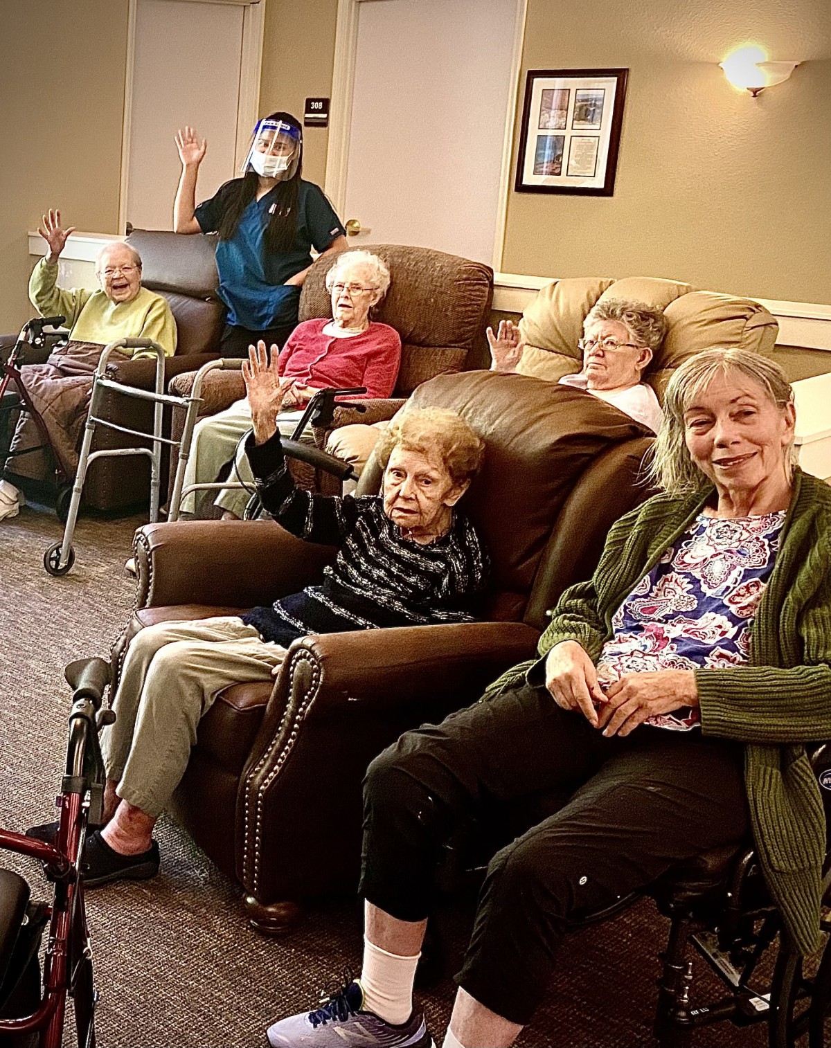 A group of seniors sitting together indoors, some in wheelchairs, smiling and waving.