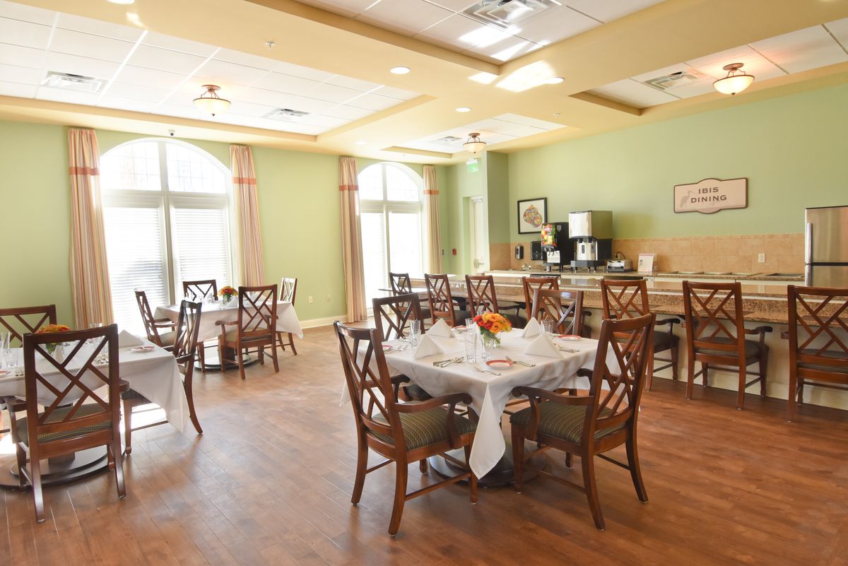 A well-lit dining room with neatly arranged tables and chairs, featuring large windows and wooden flooring.