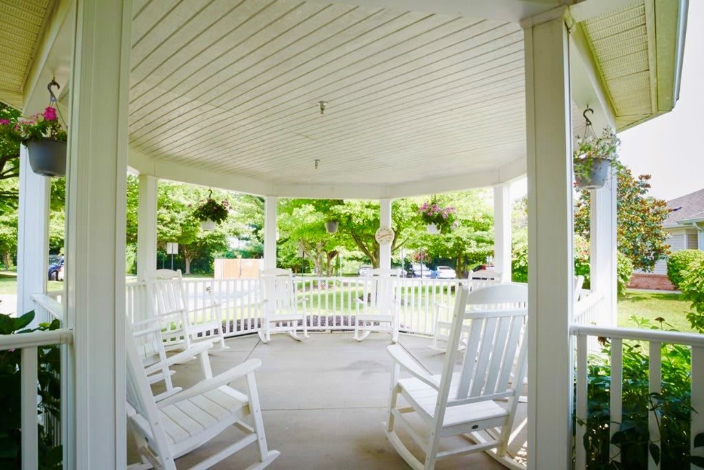A cozy outdoor gazebo with white rocking chairs and hanging plants, surrounded by lush greenery.