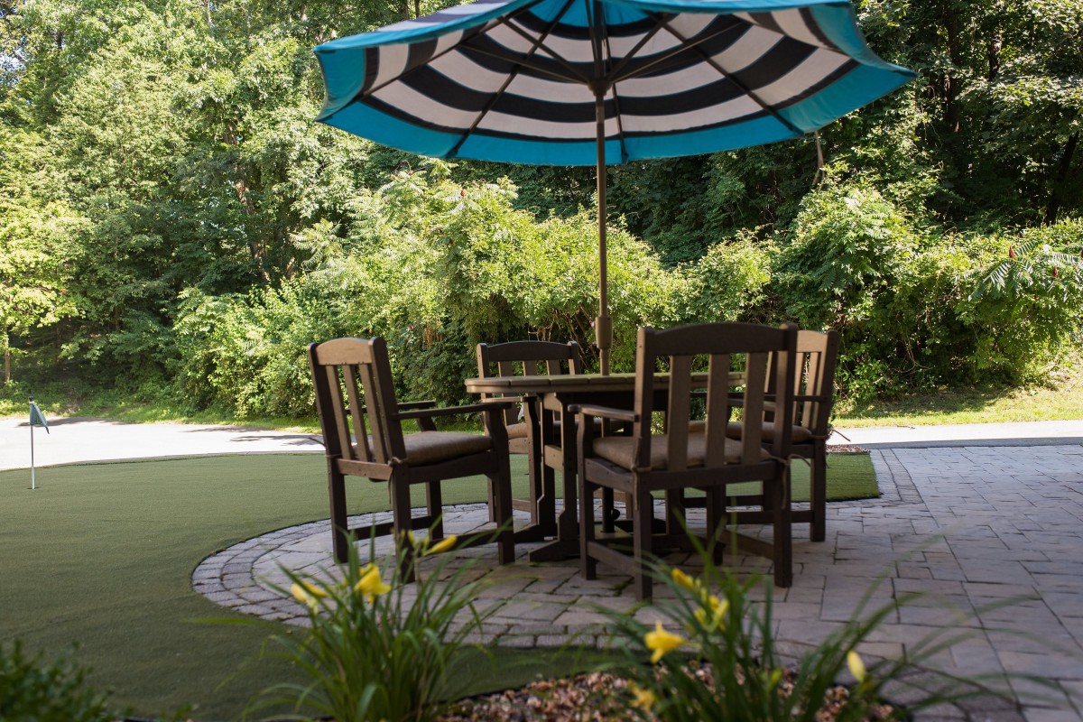 A cozy outdoor patio setup with a table and chairs under a large striped umbrella, surrounded by greenery.