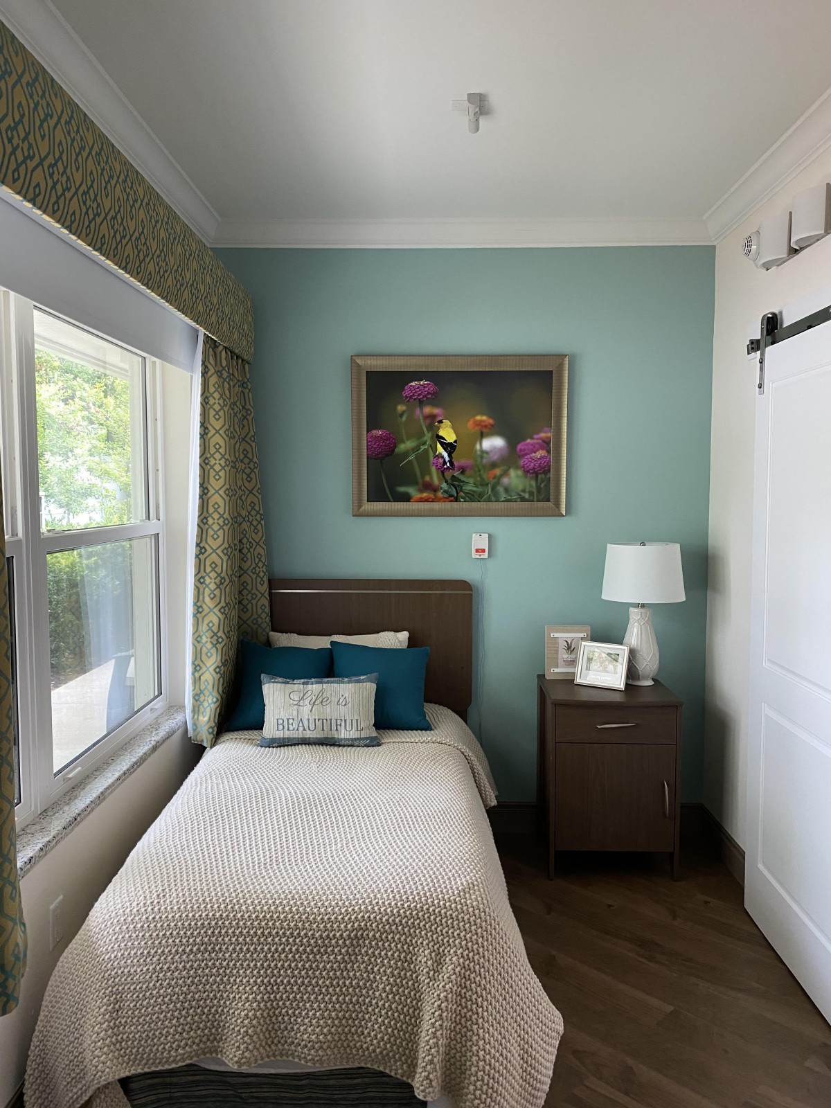 A cozy bedroom with a single bed, nightstand, and a lamp by the window.