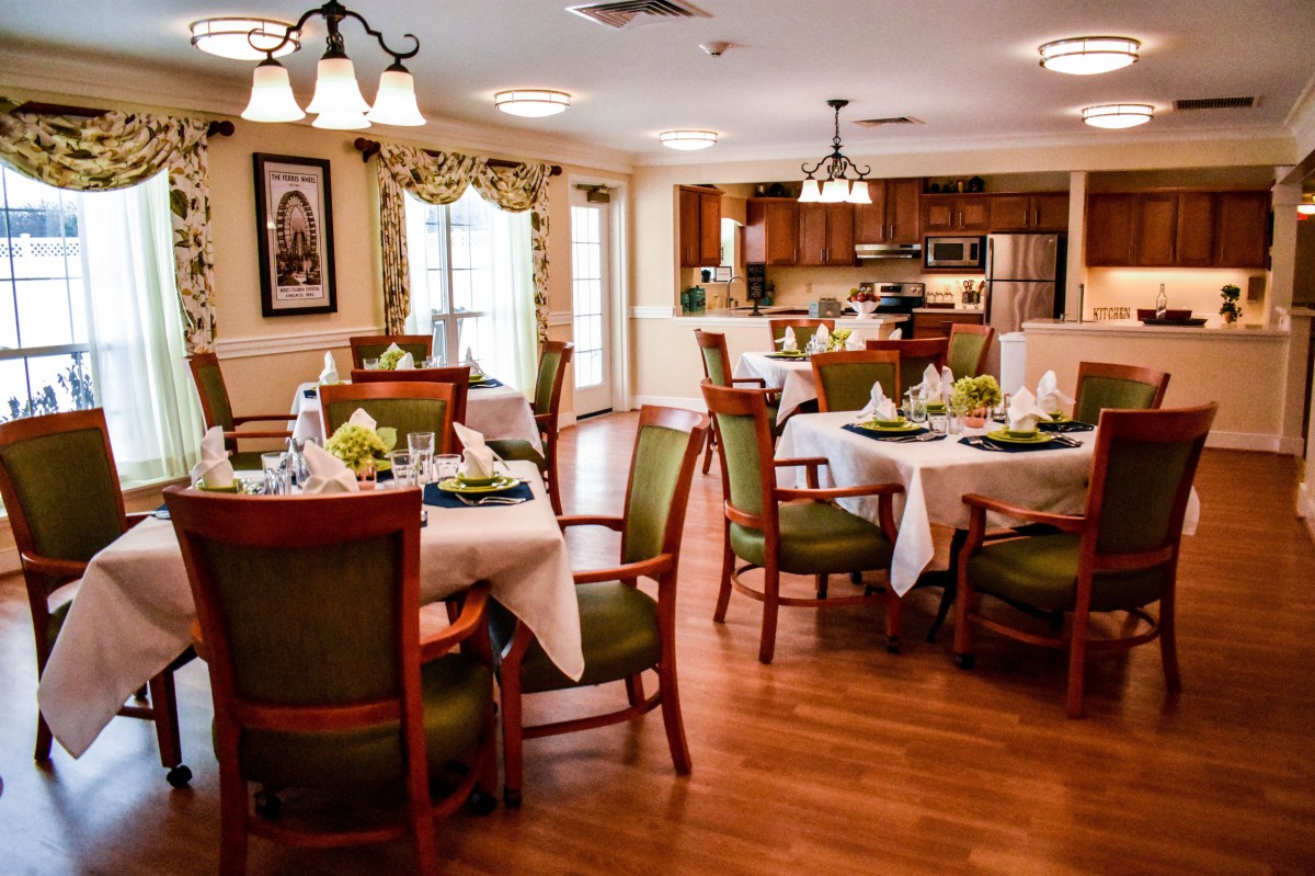 A warmly decorated dining room with neatly set tables and comfortable chairs, featuring a view of the kitchen area.