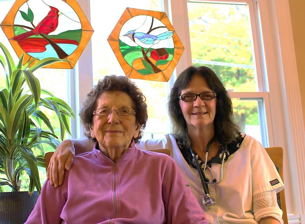 An elderly woman and a younger woman sitting together in front of a window with stained glass art.