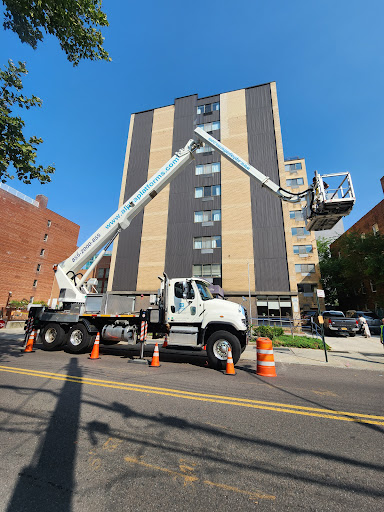 A bucket truck is extended towards a high-rise building for maintenance work, surrounded by traffic cones in an urban setting.