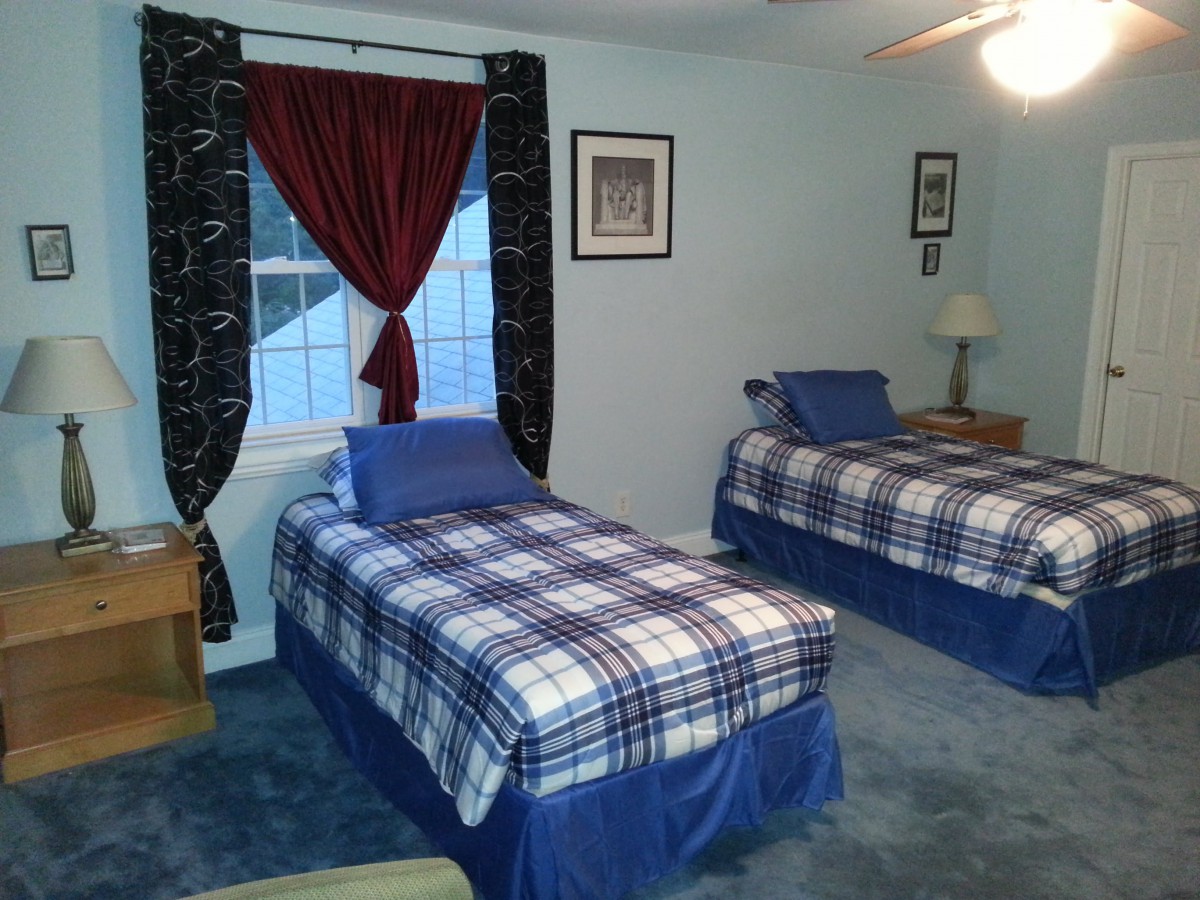 A cozy bedroom featuring two twin beds with blue plaid bedding, a carpeted floor, and a window adorned with red and black curtains.