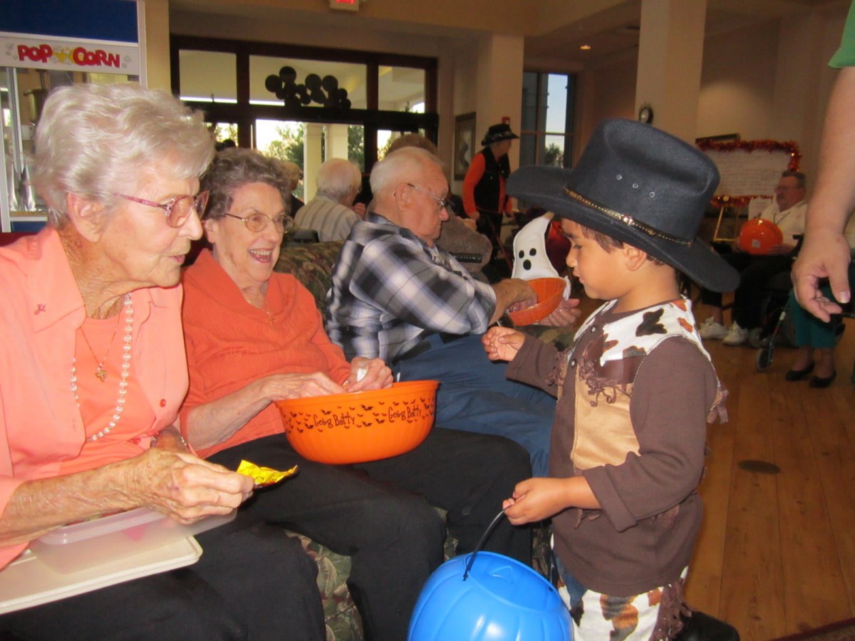 A child in a Halloween costume interacts with elderly women holding candy bowls indoors.