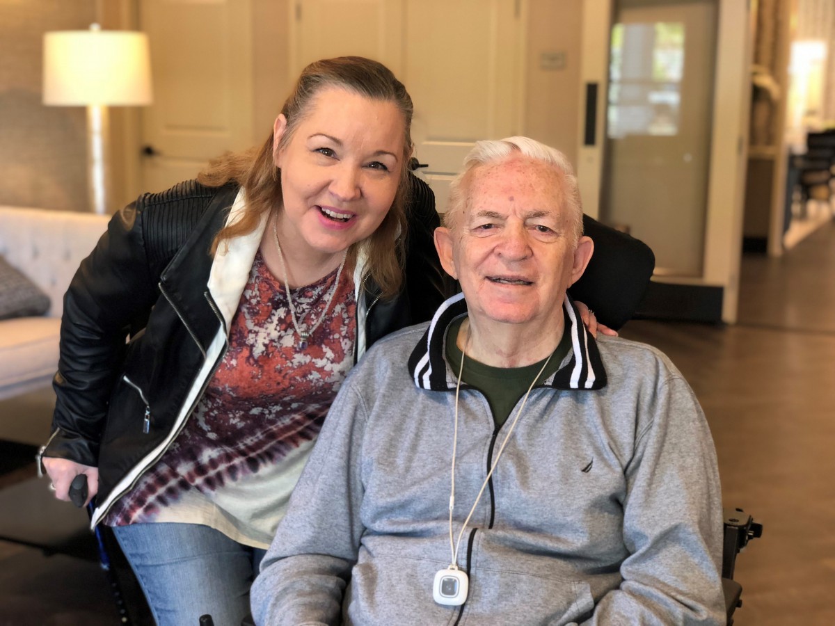 A woman smiling while standing next to an elderly man seated indoors.