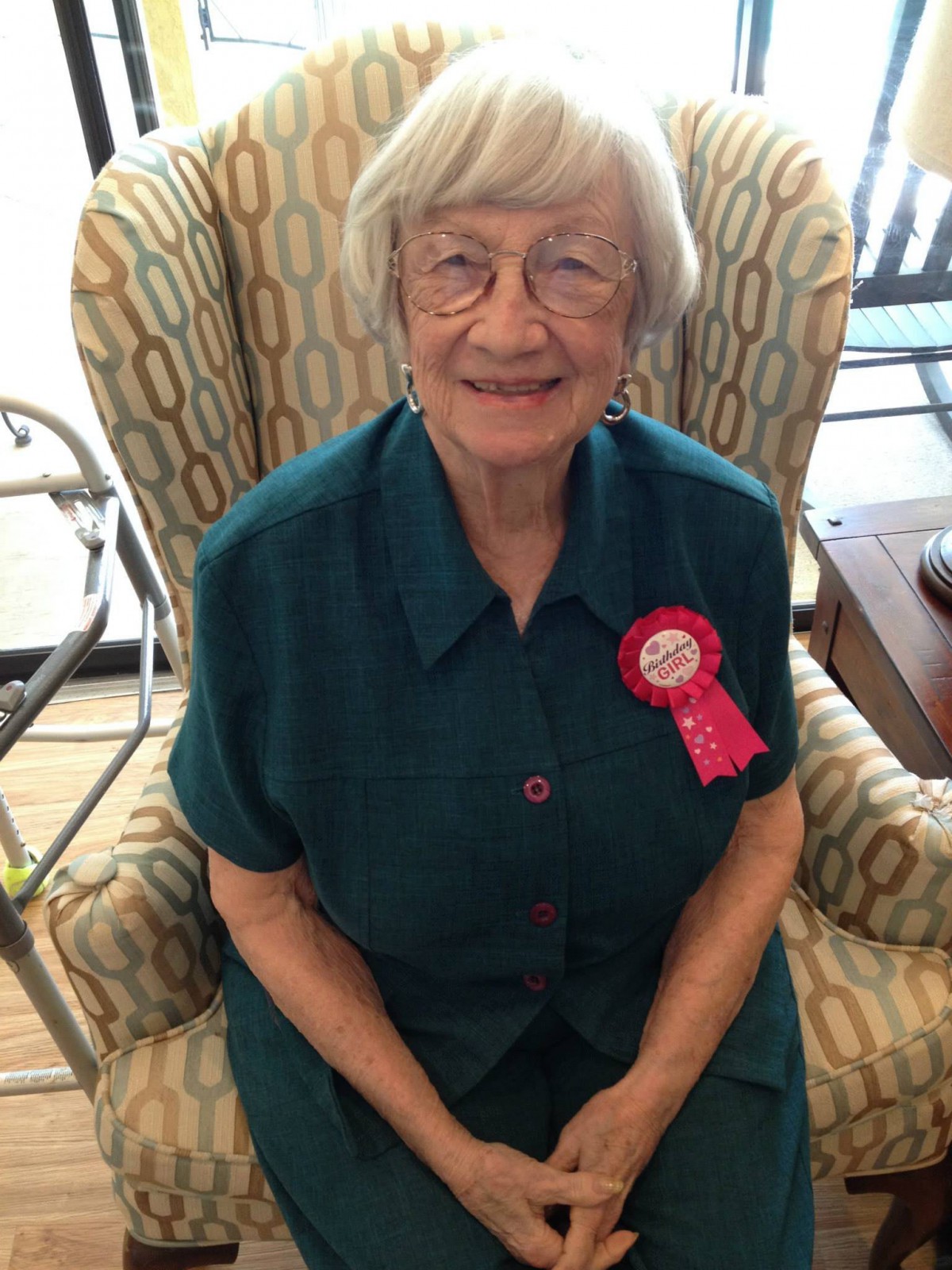 An elderly woman smiling while seated in an armchair, wearing a birthday ribbon.