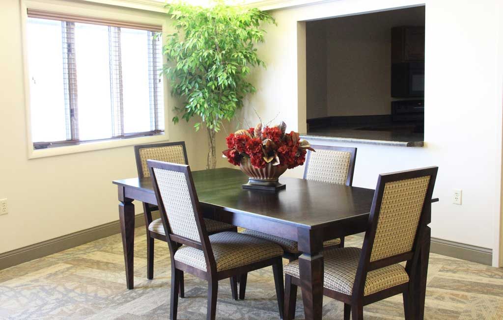 A modern dining room with a dark wooden table, upholstered chairs, and a decorative centerpiece of red flowers, complemented by a green plant in the corner.