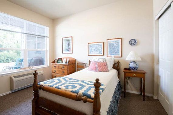 A cozy bedroom with a single wooden bed, adorned with a patterned quilt and decorative pillows. The room features warm lighting, a small nightstand with a lamp, and framed artwork on the walls.
