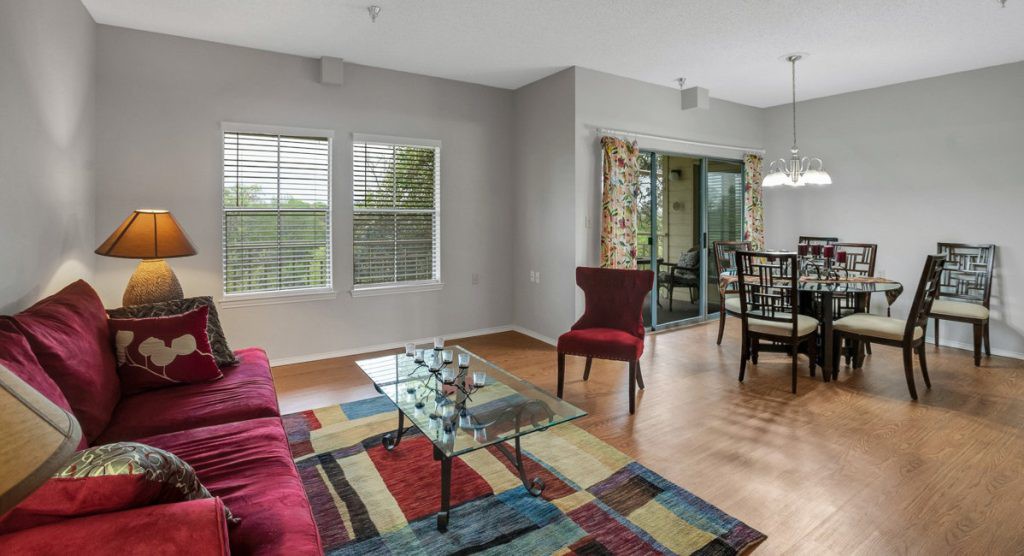 A modern living room with a colorful rug, red chair, and a glass coffee table, adjacent to a dining area with large windows.