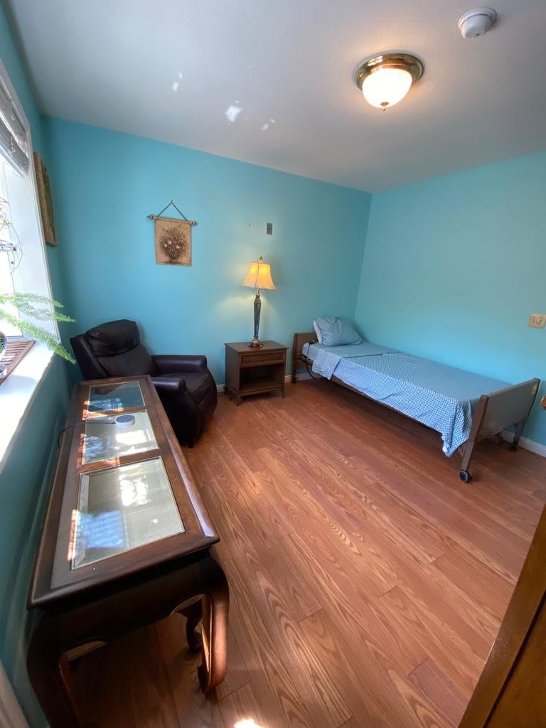 A small bedroom with blue walls features a single bed, armchair, lamp, and a window letting in natural light.