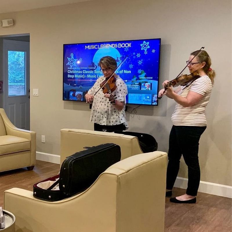 Two musicians performing indoors, playing violins in a living room setting.