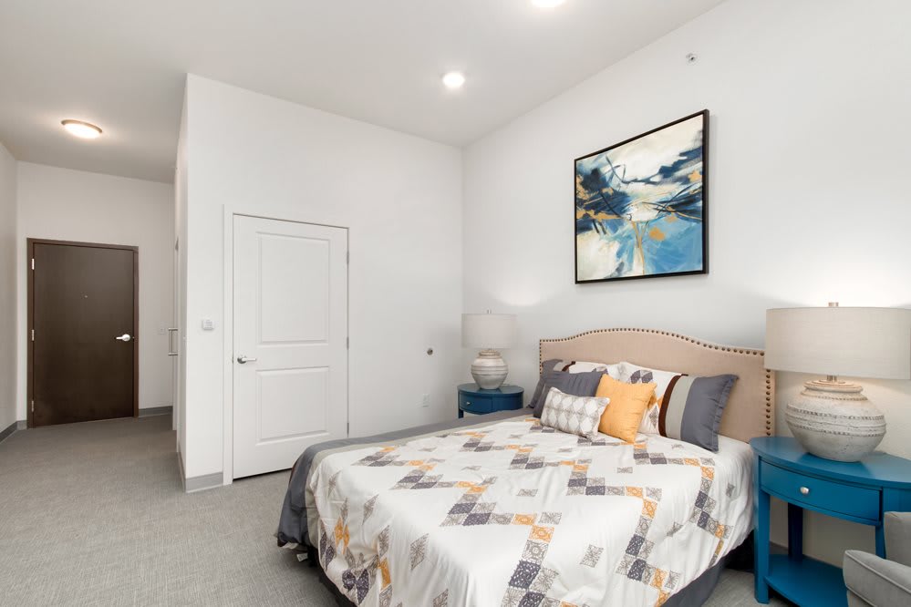 A bright and minimalist bedroom featuring a neatly made bed with patterned bedding, a bedside lamp, and abstract artwork on the wall.