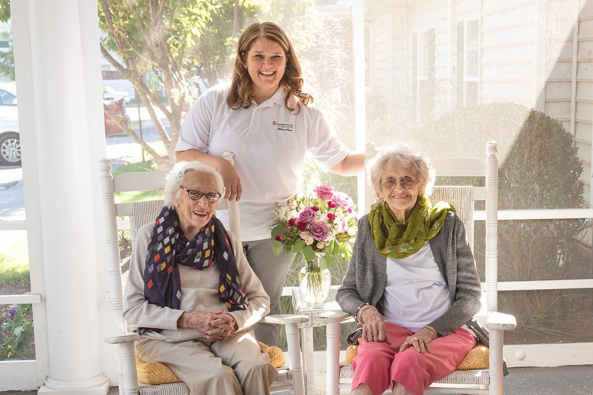 Two elderly women sit on a porch with a caregiver standing behind them, surrounded by flowers.