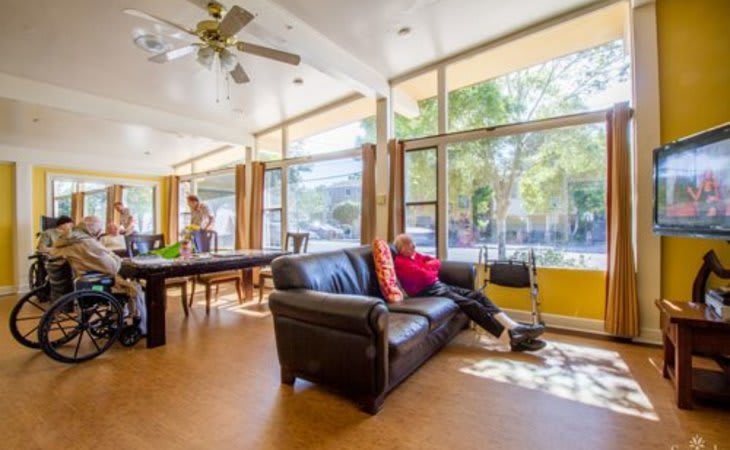 A brightly lit living room with large windows, featuring a comfortable sofa and a ceiling fan.