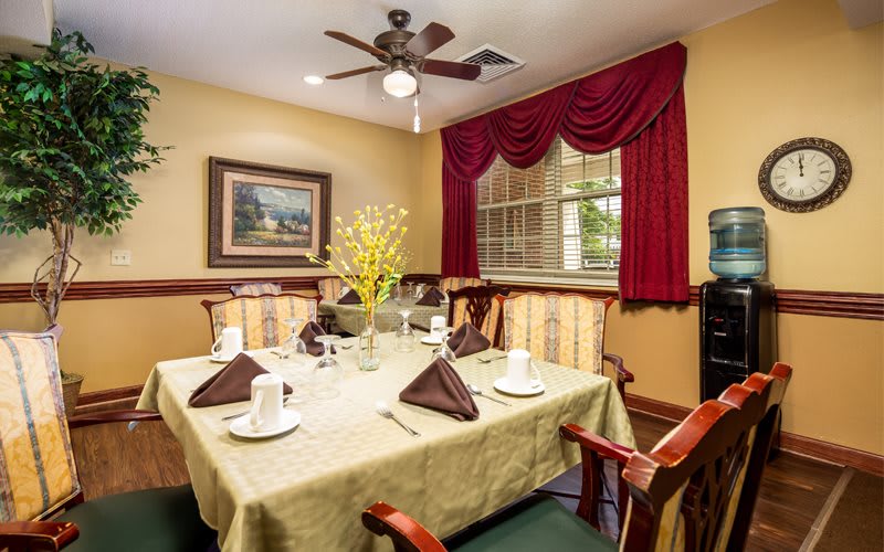 An elegantly set dining table in a cozy dining room with a ceiling fan and red drapes.