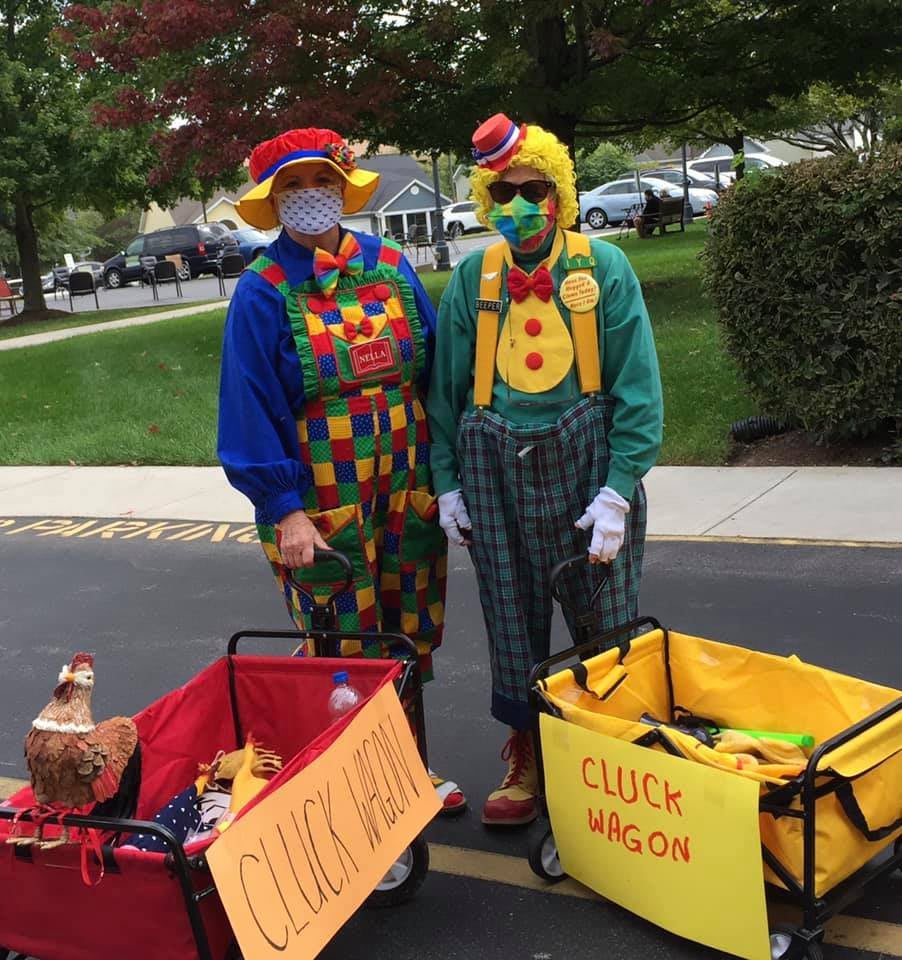 Two clowns in colorful costumes and masks stand with wagons in a park.